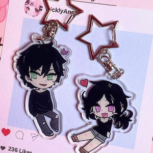 Puede incluir: Dos llaveros de acrílico con personajes estilo anime de cabello negro, cada uno unido a un broche plateado en forma de estrella. Un personaje tiene ojos verdes y viste un suéter negro, mientras que el otro tiene ojos rosas y un top negro.