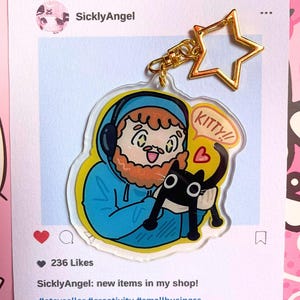 Könnte beinhalten: Ein klarer Acryl-Schlüsselanhänger mit einer Cartoon-Illustration einer Person mit blauem Kapuzenpullover und Kopfhörern, die eine schwarze Katze hält. Der Schlüsselanhänger hat einen goldfarbenen sternförmigen Verschluss. Der Text auf dem Bild lautet "KITTY!!".