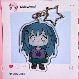 May include: A clear acrylic keychain with a cartoon girl design. The girl has blue hair, a purple top with a paw print, and a green skirt. The keychain includes a silver star charm and a metal clasp. The background is a pink and white Instagram-themed card.