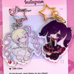 May include: Two anime-style keychains featuring a light-haired angel and a dark-haired girl in a black and red dress. Each keychain has a star-shaped charm. The background is a pink Instagram-themed graphic with text.