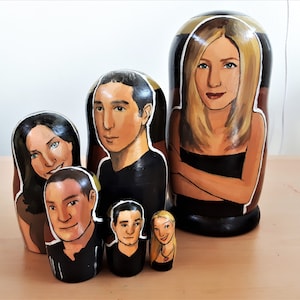 Peut inclure: Un ensemble de six poupées gigognes, chacune peinte avec un portrait différent d'une personne. La plus grande poupée est peinte avec une femme aux longs cheveux blonds, la suivante est un homme aux cheveux foncés, la suivante est une femme aux cheveux foncés, la suivante est un homme aux cheveux courts foncés, la suivante est un homme aux cheveux courts foncés, et la plus petite est une femme aux cheveux blonds.