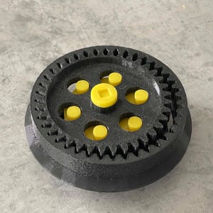 Könnte beinhalten: Ein schwarz-gelber 3D-gedruckter Getriebemechanismus. Das Zahnrad hat ein kreisförmiges Design mit Zähnen am äußeren Rand und mehreren gelben zylindrischen Stiften. Die Mitte hat einen gelben quadratischen Stift.