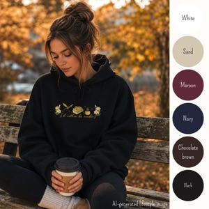 Könnte beinhalten: Schwarzer Kapuzenpullover mit goldenem Blumen- und Buchdesign, Text "I'd rather be reading". Der Hoodie wird von einer Person getragen, die auf einer Holzbank sitzt. Der Hintergrund zeigt Herbstlaub und eine Farbpalette aus Weiß, Sand, Burgunderrot, Marineblau, Braun und Schwarz.