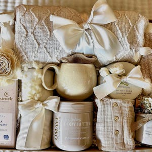 May include: A gift basket filled with cream-colored items, including a blanket, socks, a mug, a jar of body butter, a candle, a bar of milk chocolate, and a baby outfit. The items are tied with white ribbons and arranged in a wooden box.