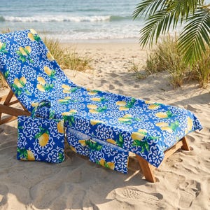 Op de afbeelding: Een blauw-wit strandlaken en een bijpassende tas met een herhalend patroon van gele citroenen en groene bladeren. Het laken ligt over een houten ligstoel gedrapeerd, terwijl de tas ernaast in het zand staat.