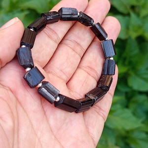 Könnte beinhalten: Ein Armband aus schwarzem Turmalin mit rechteckigen Perlen und silberfarbenen Abstandshaltern. Das Armband wird in einer Hand vor einem Hintergrund aus grünen Blättern präsentiert. Die Perlen haben ein natürliches, unpoliertes Aussehen.