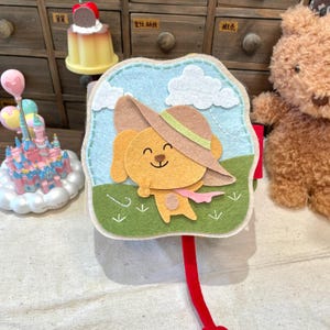 Puede incluir: Un marcapáginas de fieltro con un perro de dibujos animados sonriente con un sombrero, sobre un cielo azul con nubes blancas y hierba verde. El marcapáginas tiene una cinta roja adjunta. El diseño es caprichoso y colorido.
