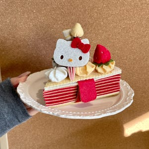 Puede incluir: Una porción de pastel de fieltro con una cara de Hello Kitty blanca y un lazo rojo, una fresa roja y decoraciones de merengue. El pastel tiene capas a rayas rojas y blancas y se presenta en un plato blanco.