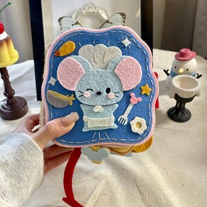 Puede incluir: Un libro de actividades de fieltro con una cubierta azul que presenta un ratón chef. El ratón tiene orejas rosas y un gorro y delantal blancos. El libro tiene varios alimentos de fieltro y una lengüeta roja.