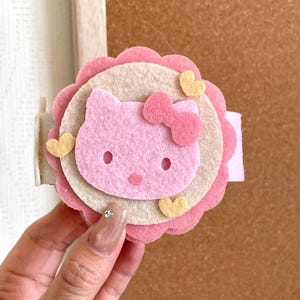 Puede incluir: Pinza para el pelo de fieltro rosa y blanco con la cara de un gato de dibujos animados con un lazo. La pinza tiene un diseño en capas con un borde festoneado y pequeños acentos de corazón amarillo. El diseño general es lindo y juguetón.