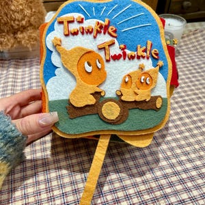 Puede incluir: Un libro de fieltro hecho a mano con las palabras "Twinkle Twinkle" en letras rojas. El libro presenta personajes naranjas y un fondo de cielo azul. El libro tiene un asa amarilla.