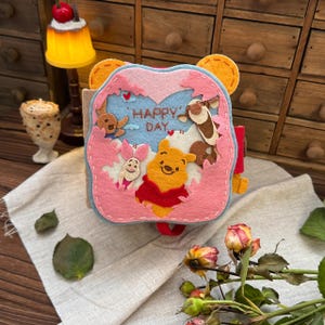 Puede incluir: Un libro de fieltro hecho a mano con Winnie the Pooh y sus amigos. La portada es rosa y azul con las palabras "Happy Day". El libro tiene una correa roja y orejas de oso amarillas en la parte superior. El fondo incluye una lámpara y flores.