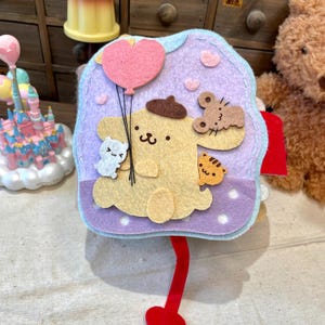 Puede incluir: Una manualidad de fieltro con un personaje de dibujos animados amarillo sosteniendo un globo con forma de corazón rosa. El personaje está rodeado de otros recortes de fieltro, incluyendo un ratón, un gato y una pequeña criatura blanca. El fondo es lila claro.