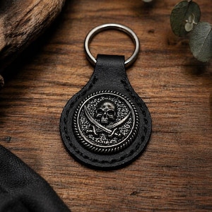 May include: Black leather keyring featuring a silver skull and crossbones emblem. The emblem is circular and intricately designed. The keyring has a silver ring for keys. Set against a wooden background.