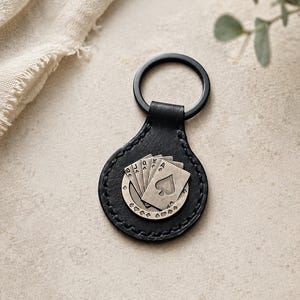 May include: A black leather circular keyring featuring a metallic silver-toned emblem of a royal flush poker hand. The emblem displays five playing cards with the ace of spades in front, surrounded by a border of small heart symbols.