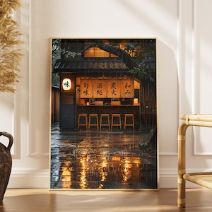 May include: Framed print of a Japanese restaurant in the rain. The restaurant has a wooden facade, lit interior, and a row of stools. The wet street reflects the warm light. Japanese characters are visible on the signage.