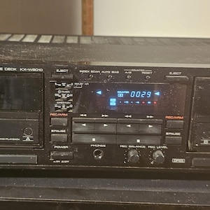May include: A black Kenwood stereo double cassette deck model KX-W8010 featuring two cassette compartments, a central digital display showing 0029, various control buttons, playback transport keys, and two rotary knobs for recording levels.