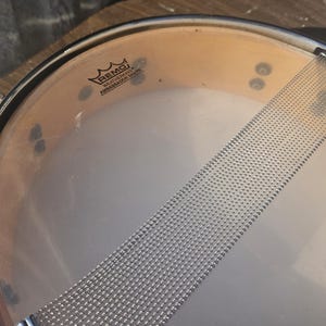 May include: Close-up of a snare drum. The drumhead is transparent with a silver snare wire stretched across the bottom. The drum's rim is black, and the shell is a light brown color. The Remo logo is visible on the drumhead.