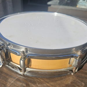 May include: A snare drum with a white drumhead and a light brown wooden body. The drum has a silver-colored metal rim and hardware. The drum is sitting on a wooden surface.