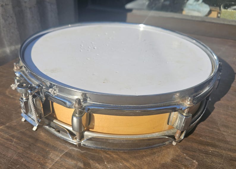 May include: A snare drum with a white drumhead and a light brown wooden body. The drum has a silver-colored metal rim and hardware. The drum is sitting on a wooden surface.