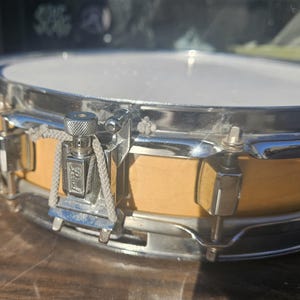 May include: Close-up of a snare drum with a light-colored wooden body and a white drumhead. The drum has chrome hardware, including tension rods and a Pearl logo. The drum is designed for musical performances.