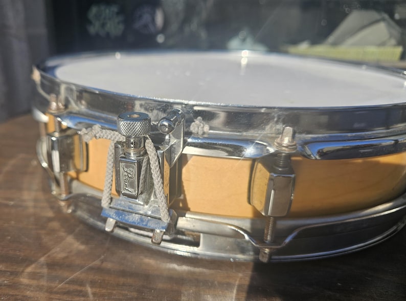 May include: Close-up of a snare drum with a light-colored wooden body and a white drumhead. The drum has chrome hardware, including tension rods and a Pearl logo. The drum is designed for musical performances.