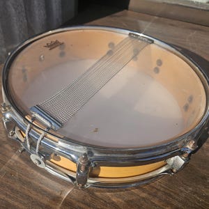 May include: A vintage snare drum with a clear drumhead and a chrome-plated rim. The drum has a natural wood finish on the shell and a wire snare. The drum is designed for percussion instruments.
