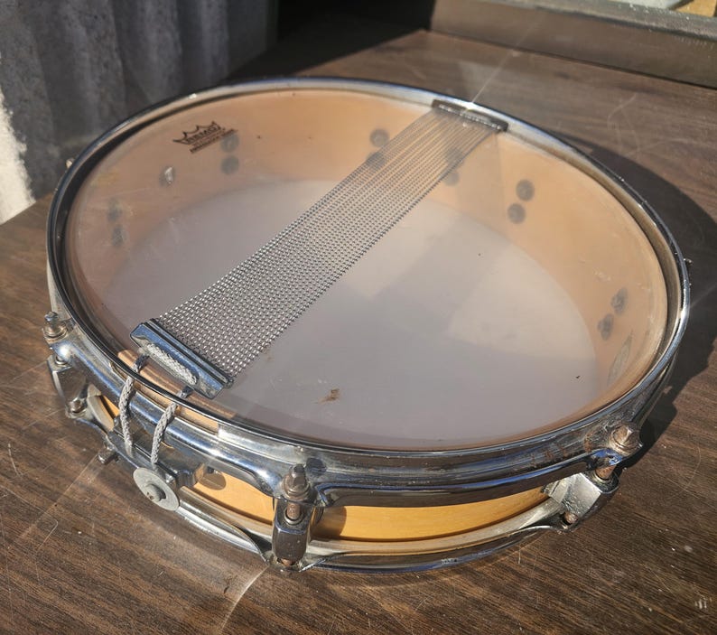 May include: A vintage snare drum with a clear drumhead and a chrome-plated rim. The drum has a natural wood finish on the shell and a wire snare. The drum is designed for percussion instruments.