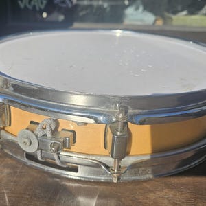 May include: A snare drum with a light-colored wooden shell and a white drumhead. The drum has a chrome-plated rim and hardware. The drum is sitting on a wooden surface. The drum is a musical instrument.
