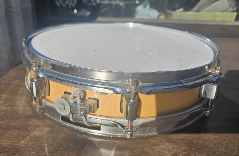 May include: A snare drum with a light-colored wooden shell and a white drumhead. The drum has a chrome-plated rim and hardware. The drum is sitting on a wooden surface. The drum is a musical instrument.