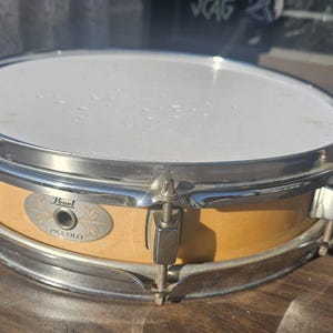 May include: A Pearl Piccolo snare drum with a light wood finish and chrome hardware. The drum has a white drumhead and is designed for a high-pitched sound. The drum is sitting on a wooden surface.