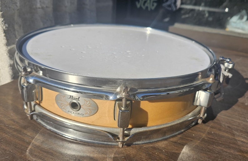 May include: A Pearl Piccolo snare drum with a light wood finish and chrome hardware. The drum has a white drumhead and is designed for a high-pitched sound. The drum is sitting on a wooden surface.