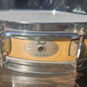 May include: A Pearl Piccolo snare drum with a light wood finish and chrome hardware. The drum features a white drumhead and a badge with the Pearl logo and the word "Piccolo".