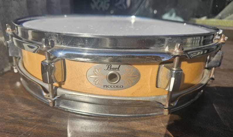 May include: A Pearl Piccolo snare drum with a light wood finish and chrome hardware. The drum features a white drumhead and a badge with the Pearl logo and the word "Piccolo".