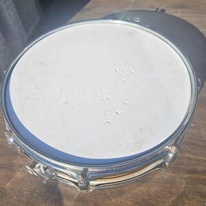 May include: A silver snare drum with a white drumhead. The drum has a shiny metal rim and hardware. The drum is sitting on a wooden surface.