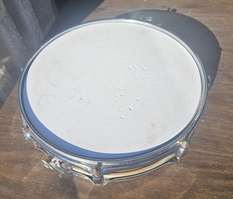 May include: A silver snare drum with a white drumhead. The drum has a shiny metal rim and hardware. The drum is sitting on a wooden surface.