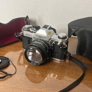 May include: A silver and black Canon AE-1 film camera with a lens, lens cap, and strap. The camera is accompanied by a black leather case and a black camera case with a red interior. The camera is on a brown surface.