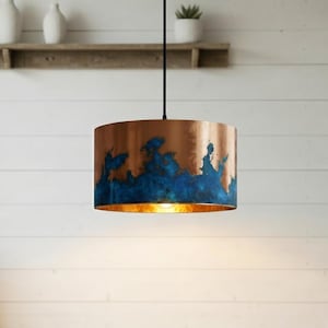 Op de afbeelding: Een koperen hanglamp met een blauwe en koperen afwerking. De cilindrische lampenkap heeft een textuur, abstract ontwerp. Het licht hangt aan een zwart snoer en verlicht de ruimte eronder. De armatuur is een decoratief element.