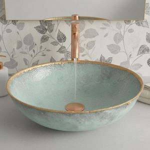 May include: A light blue, textured ceramic sink with a gold rim and a copper tap. A white soap dispenser with a copper pump and folded white towels are next to the sink. The background features a floral wallpaper.