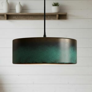 Może przedstawiać: Cylindryczna lampa wisząca z gradientowym wykończeniem, przechodzącym od ciemnobrązowego do turkusowego. Lampa wisi na czarnym kablu. Tło: biała ściana i drewniana półka z dekoracjami.