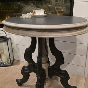 May include: A round accent table with a dark grey top and ornate black legs. The table has a cream-coloured trim and a central support column. A teacup and decorative beads rest on the tabletop. The table is approximately 61 cm in diameter.