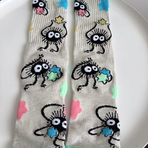 May include: A pair of beige crew socks featuring a repeating pattern of black soot sprites with large white eyes and pink, blue, yellow, and green star-shaped accents. The socks have a ribbed texture and a solid mint green cuff and toe section.