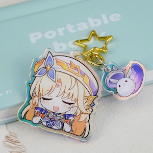 May include: An acrylic keychain featuring a blonde anime-style character with closed eyes and a blue and gold outfit, attached to a gold star-shaped clasp with a smaller secondary charm depicting a white rabbit-like creature with purple accents.