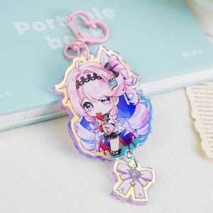 May include: An acrylic keychain featuring a chibi-style character with long pink hair, wearing a dark outfit with blue accents, attached to a pink heart-shaped clasp. A smaller charm shaped like a light purple bow with a central gem hangs below.