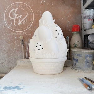 Può includere: Un contenitore bianco in ceramica a forma di cestino pasquale con una parte superiore a forma di uovo. Il contenitore ha un motivo a intreccio di cestino sul fondo ed è decorato con uova e accenti floreali. La parte superiore presenta diversi piccoli fori rotondi.