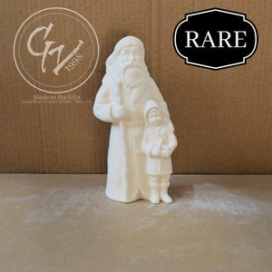 May include: A cream-colored ceramic Santa Claus figurine holding a child, with a long coat and a white beard. The figurine has the text "RARE" in a black and white label. The image also includes the text "Made in the USA".