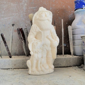 May include: A cream-colored ceramic figurine of a man with a long beard, hat, and coat, holding a staff and accompanied by a dog. The figurine is unpainted and stands on a flat base. The background includes tools and containers.