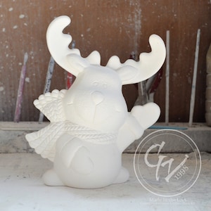 May include: White ceramic reindeer figurine with a scarf, standing on a light brown surface. The reindeer has a friendly expression and is holding its right hoof up in a waving gesture.
