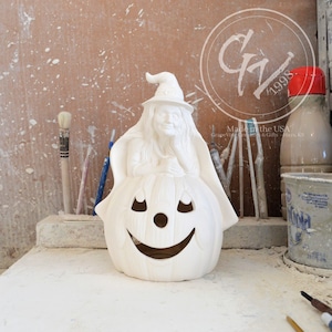 May include: White ceramic pumpkin with a witch figurine sitting inside. The witch is wearing a hat and has long hair. The pumpkin has a carved face with a smile and eyes.