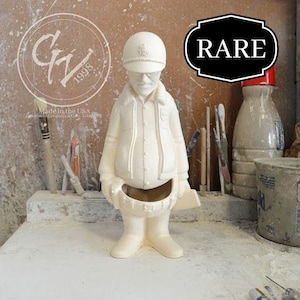 May include: White ceramic figurine of a person wearing a hat and jacket. The figurine is holding a tool in its hand. The figurine is labeled "RARE".